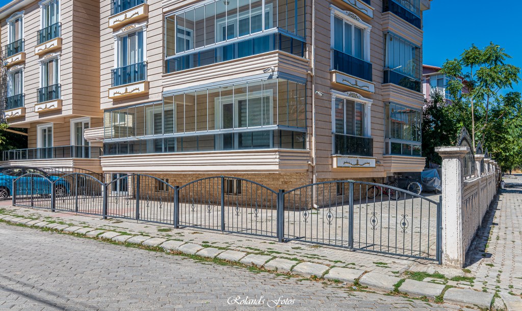Uitzicht op een modern appartementencomplex met glazen balkons en een zwart metalen hekwerk aan de voorkant, gelegen langs een straat met stenen bestrating.