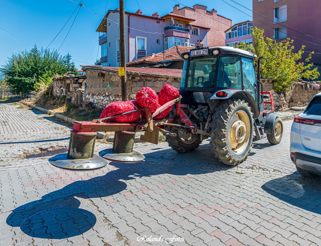 Een tractor met een rode lading rolt over een geplaveide straat met moderne gebouwen op de achtergrond.