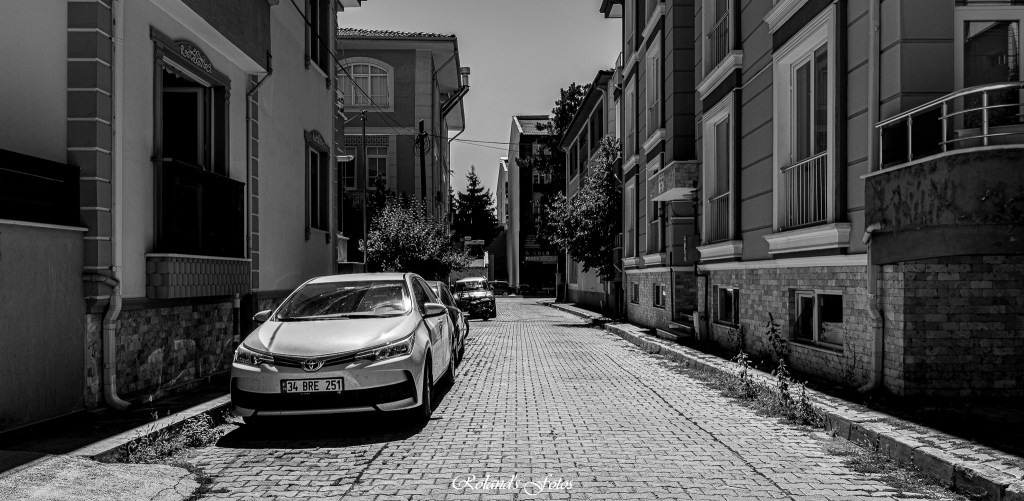 Straatbeeld in zwart-wit met een geparkeerde auto en gebouwen aan weerszijden van de smalle straat.