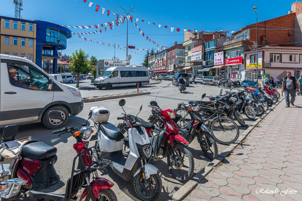 Drukke straat met verschillende voertuigen, waaronder auto's en scooters, en winkels langs de weg. Boven de straat hangen vlaggen.