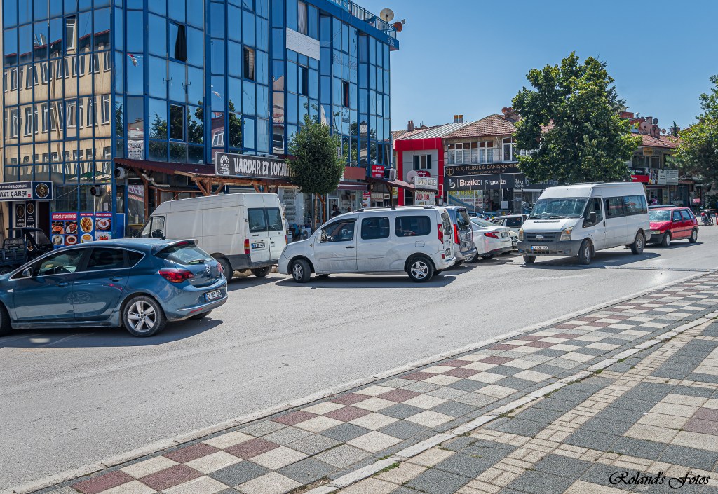 Een drukke straat met verschillende voertuigen geparkeerd en in beweging, naast moderne gebouwen met spiegelachtige gevels.