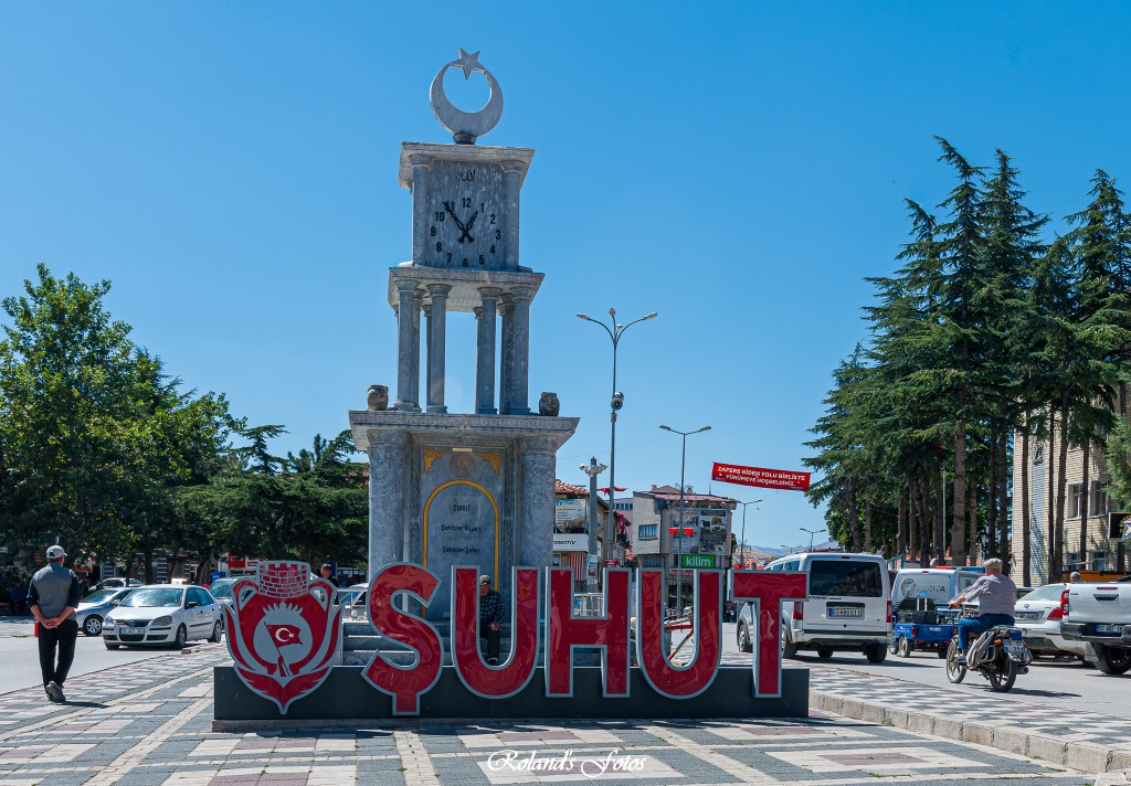 Beeld van de klok en het prominente bord 'ŞUHUT' in een stadsplein met een blauwe lucht en groene bomen op de achtergrond.