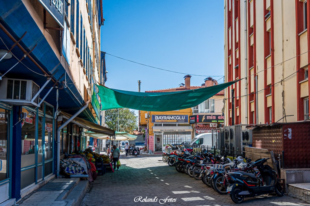 Een smalle straat met winkels en tweewielers, overschaduwd door een groene zeil, met een heldere blauwe lucht op de achtergrond.