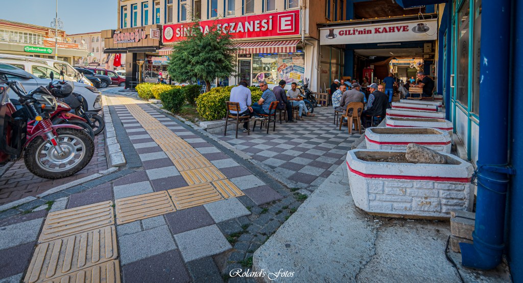 Een straat met een druk terras waar mensen zitten te genieten van koffie en elkaars gezelschap, omringd door winkels en voertuigen.