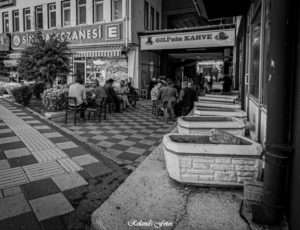 Een zwart-wit foto van een drukke straat met een café genaamd 'GİL'in KAHVE'. Mensen zitten aan tafels buiten terwijl anderen voorbij lopen.