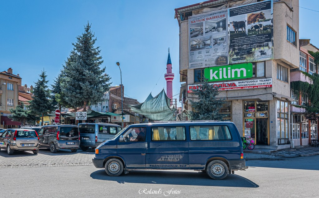 Een blauwe minibus rijdt voorbij een levendig stadsbeeld met winkels en bomen, waaronder een groot billboard met het logo 'kilim' op een gebouw.