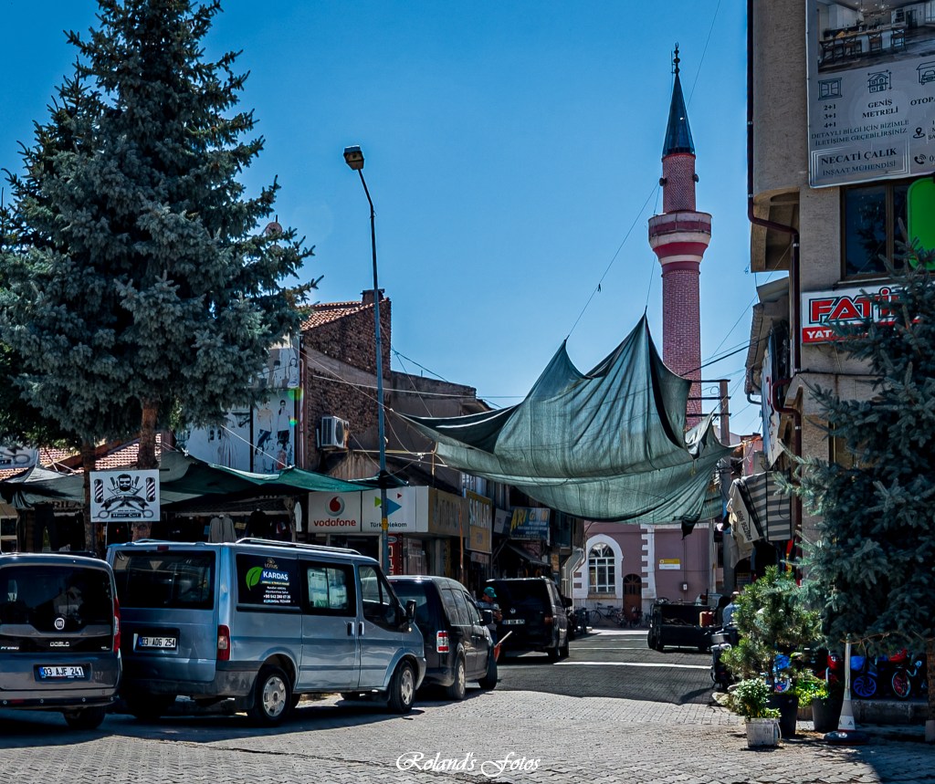 Een drukke straat met verschillende voertuigen, bomen, en een moskee met een hoge minaret, terwijl doeken over de straat zijn gespannen voor schaduw.